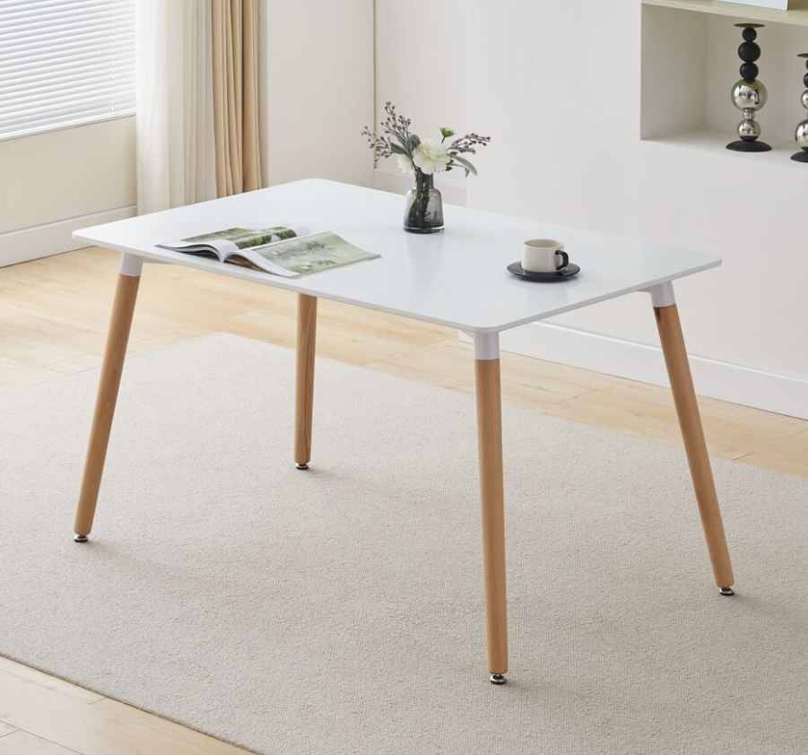 A contemporary white rectangular dining table with beech wood legs, placed on a carpeted floor in a room with an open magazine and a cup on top of it, suggesting a casual and versatile use.